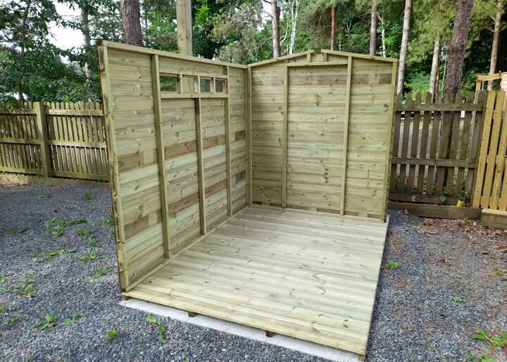 Wooden shed framework in a grassy area, surrounded by trees and a wooden fence.