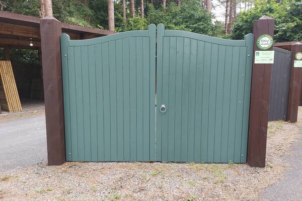 Green wooden gates with a curved top, flanked by brown posts in a wooded area.