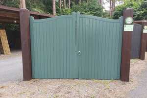 Green wooden gates with a curved top, flanked by brown posts in a wooded area.