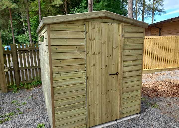 Wooden security shed with a sloped roof, situated in a gravel area with trees in the background.