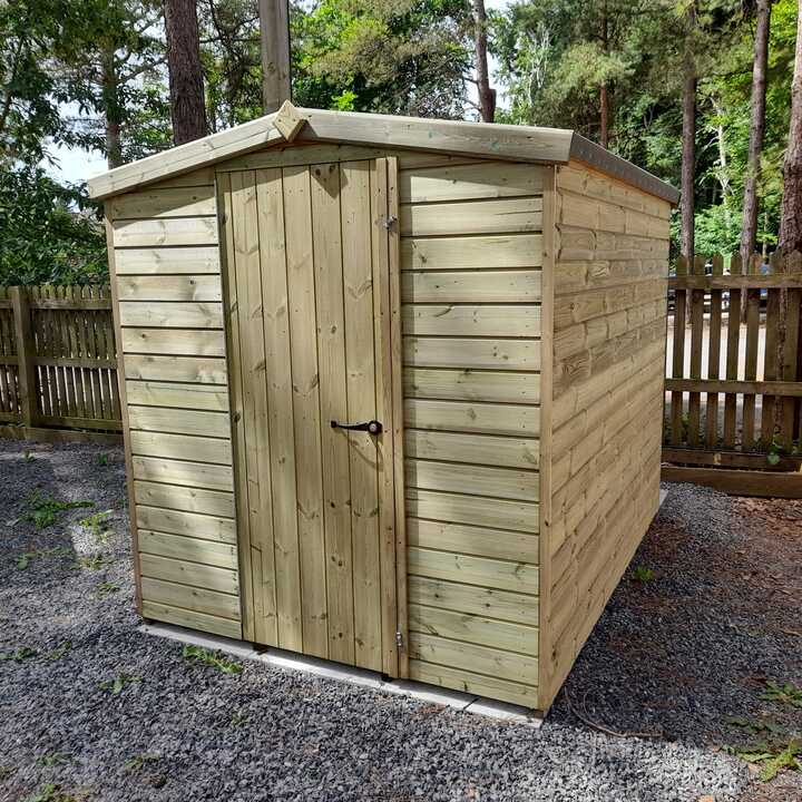 Wooden garden security shed surrounded by trees, with a gravel ground and a fenced area.