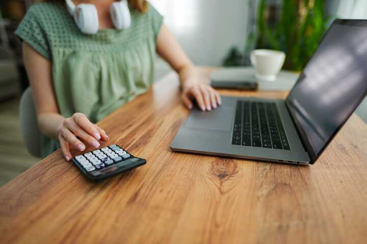 A person using a calculator next to a laptop on a wooden table in a garden office