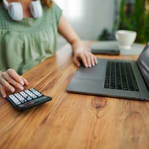 Inside a garden office a person is using a calculator beside a laptop on a wooden desk with a plant in the background.
