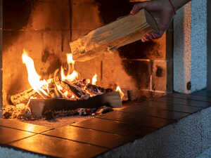 A hand places a log onto a crackling fire in a fireplace.