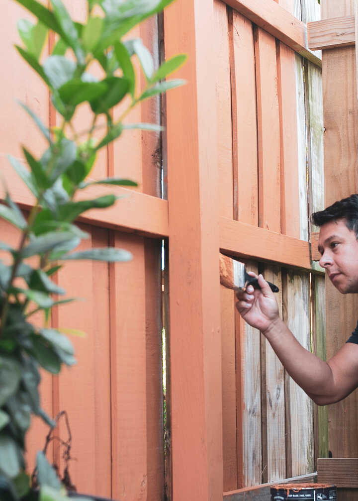 A man crouches by a fence, inspecting it closely while holding a tool.