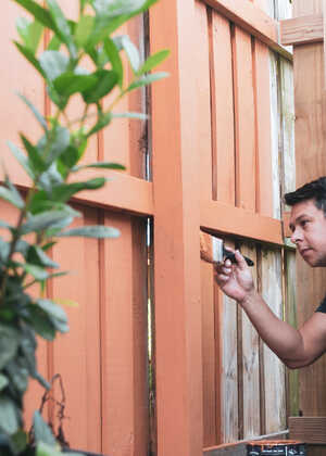 A man crouches by a fence, inspecting it closely while holding a tool.