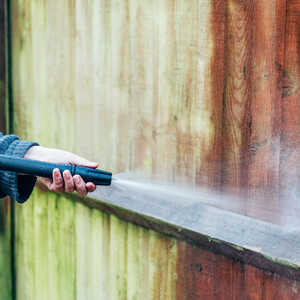 Person using a pressure washer on a wooden fence, creating steam and dirt removal.