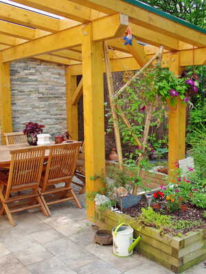 A garden pergola covers a dining table and potted plants surrounded by greenery.