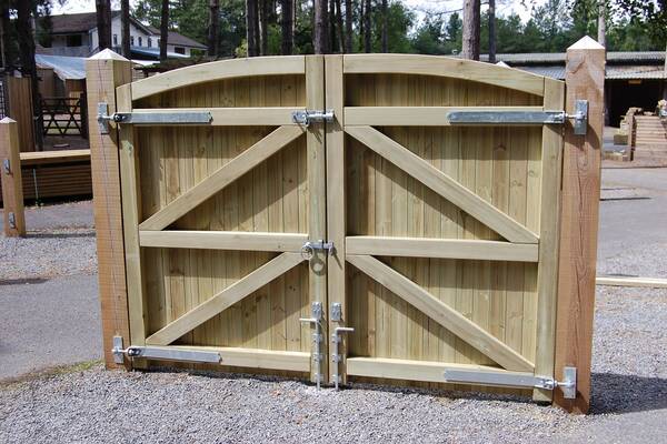 Wooden double gates with an arched top, secured with metal latches in a gravel area.