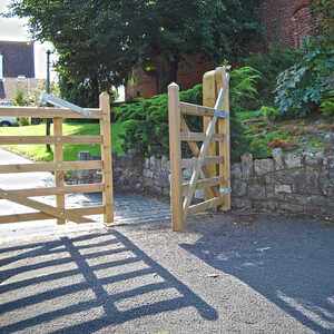 Bespoke country-style gate with pointed tops Wooden gate opening onto a path with greenery and stone wall beside it.