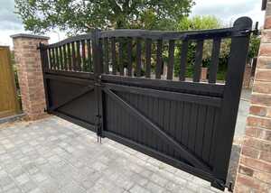 Black double gate with wooden slats, set in a brick wall with greenery in the background.
