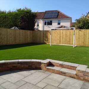 Lawn with a timber feather edge fence and a small goalpost in the background on a sunny day.