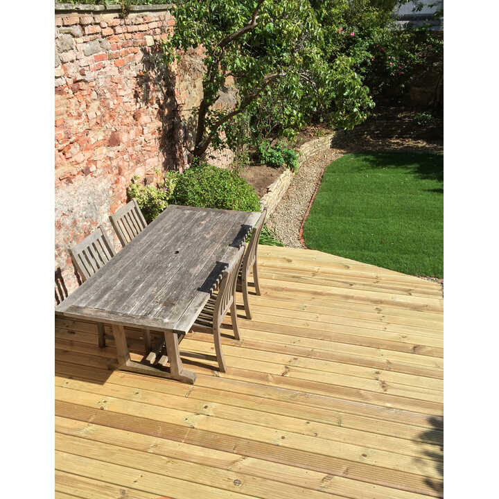 A wooden table and chairs on a wooden deck overlooking a garden with greenery.